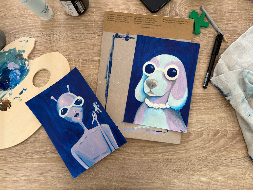 Two painted cards featuring a dog and an alien on a wooden table with art supplies.